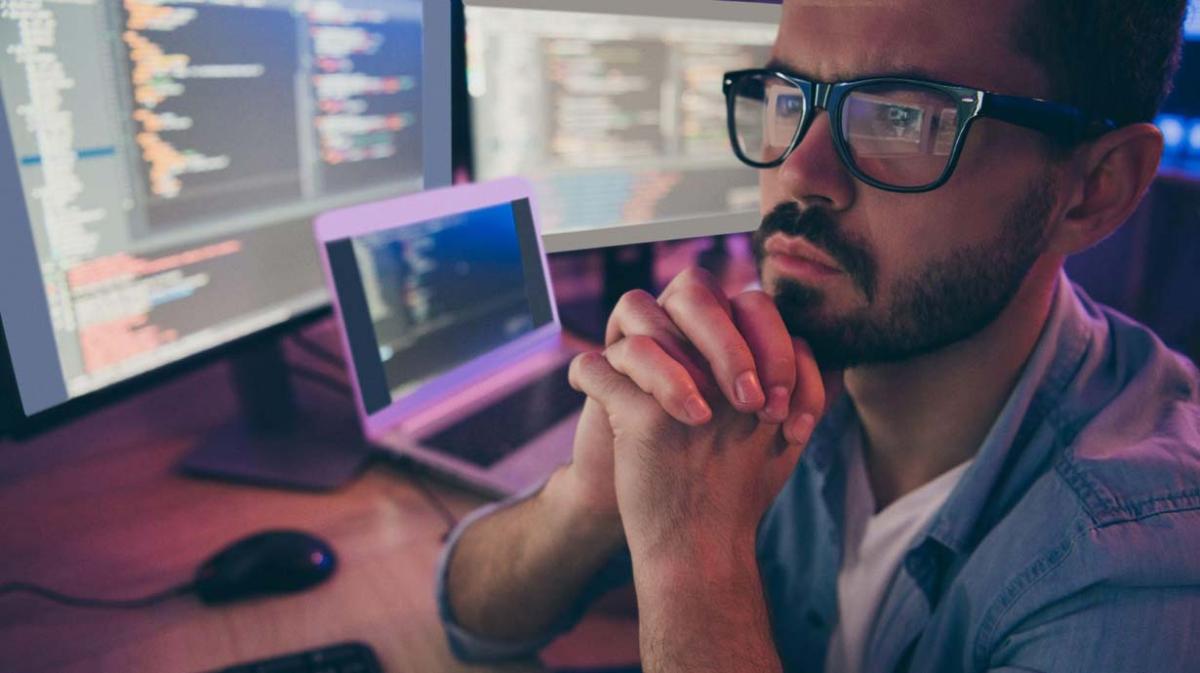 A person with folded hands looking at multiple monitors on a desk.