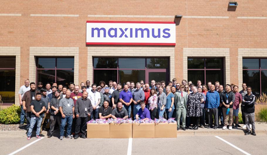 A large group posed outside a Maximus building, packed boxes in front of them.