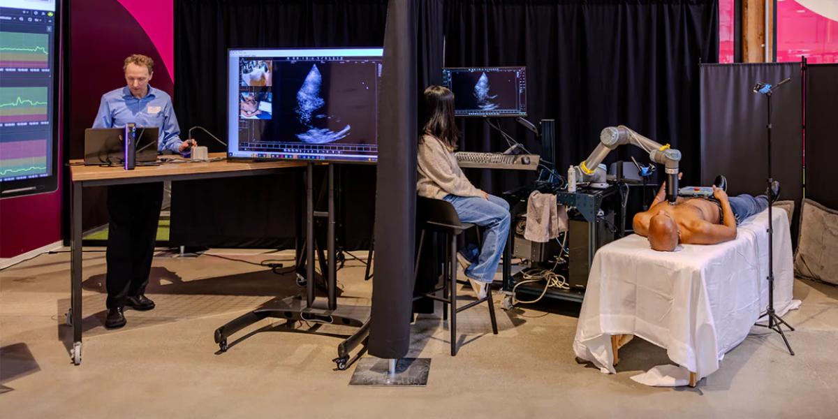 A person on a table with another sitting nearby. A medical imaging device on their chest. Another person behind a curtain viewing the images.