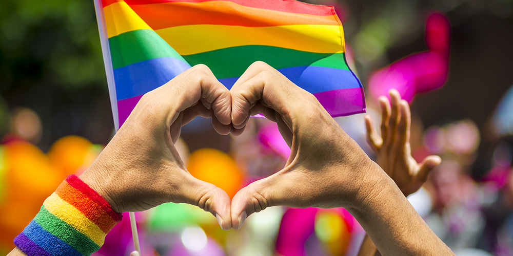 fingers forming a heart with rainbow flags in the background