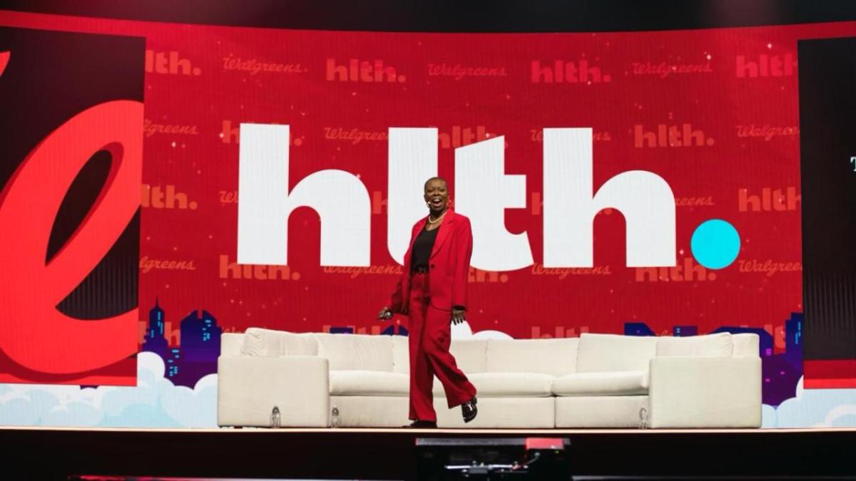 Tracey Brown on a large stage with a white couch and large digital display behind.