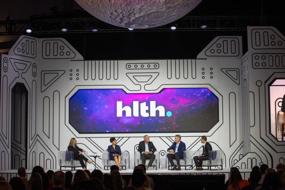 A panel of five on a stage, a digital display behind.