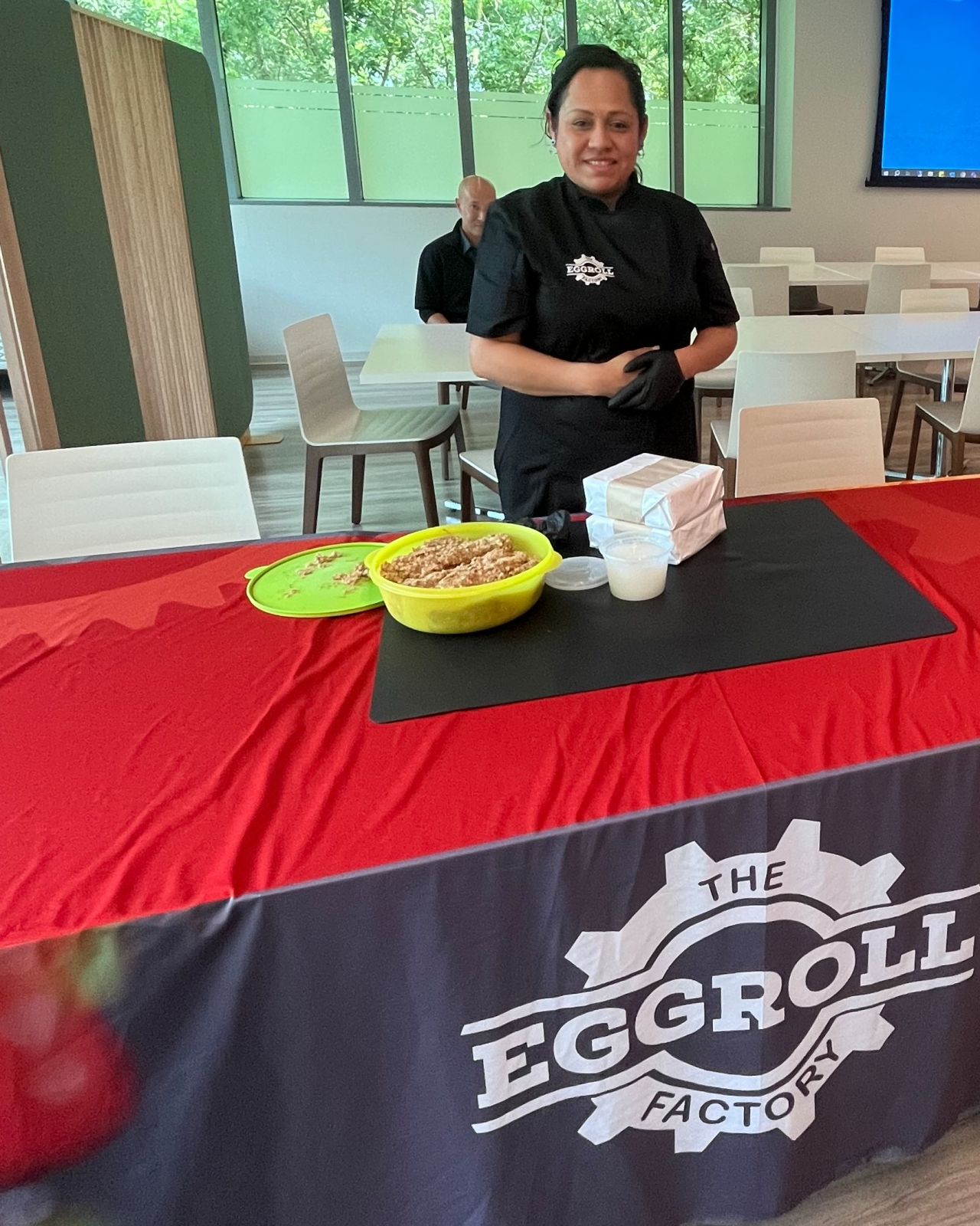A person standing with food behind a table with a cloth that says "The Eggroll Factory"