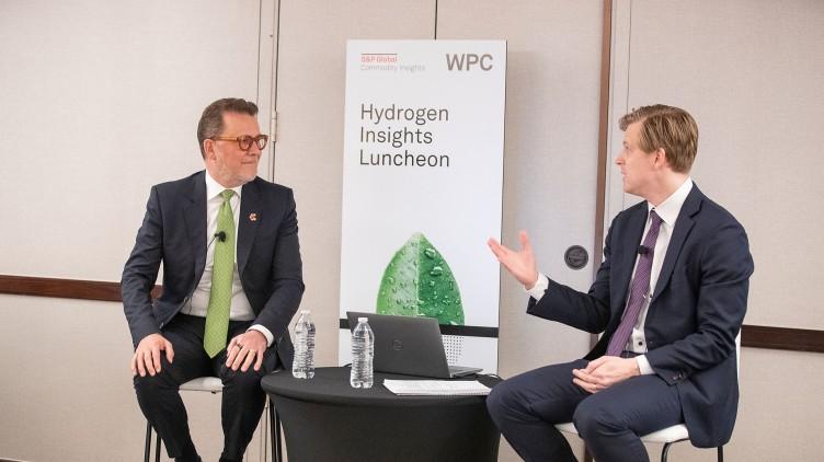 Two people sitting at a table with poster "Hydrogen Insights Luncheon" behind them