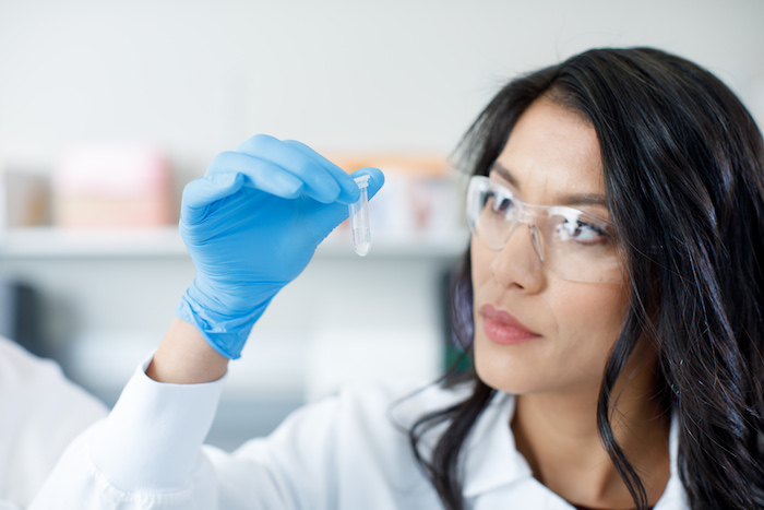 Illumina female scientist in white gown, gloves and protective eyewear looks at a small sample in a test tube.