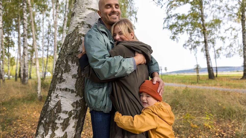 Child hugging his parents that are standing next to a tree.