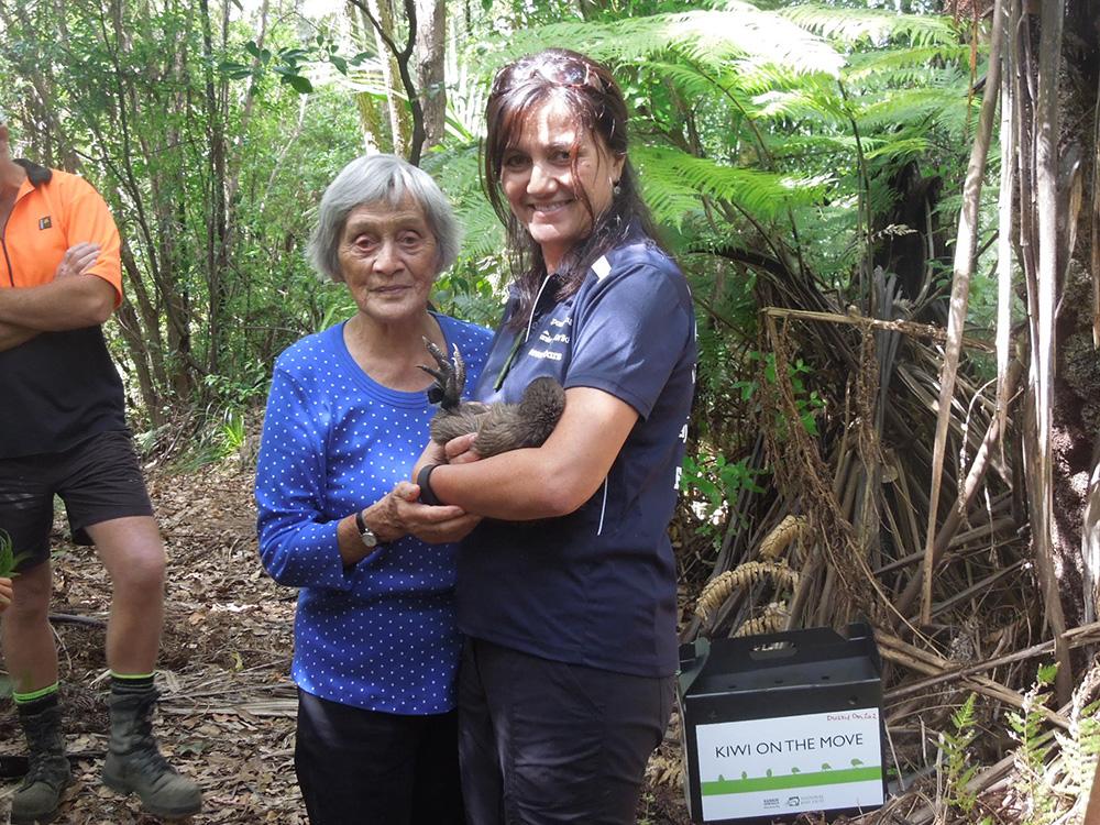 A person holding a Kiwi bird and another elder holding their arm. Both standing in a wooded area. A box with "Kiwi on the move" on the ground next to them.