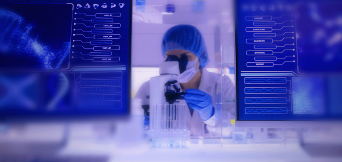 A person in medical protective wear looking in a microscope. Two computer screens to the sides.