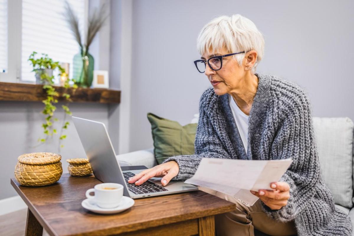 A person holding a piece of paper, looking at an open laptop in a living room setting. A tea cup next to it.