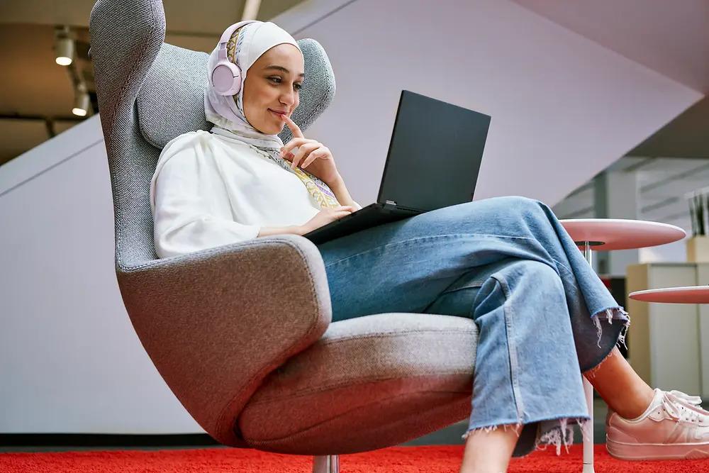 A person in a cushioned chair smiling, looking at a laptop and wearing headphones.