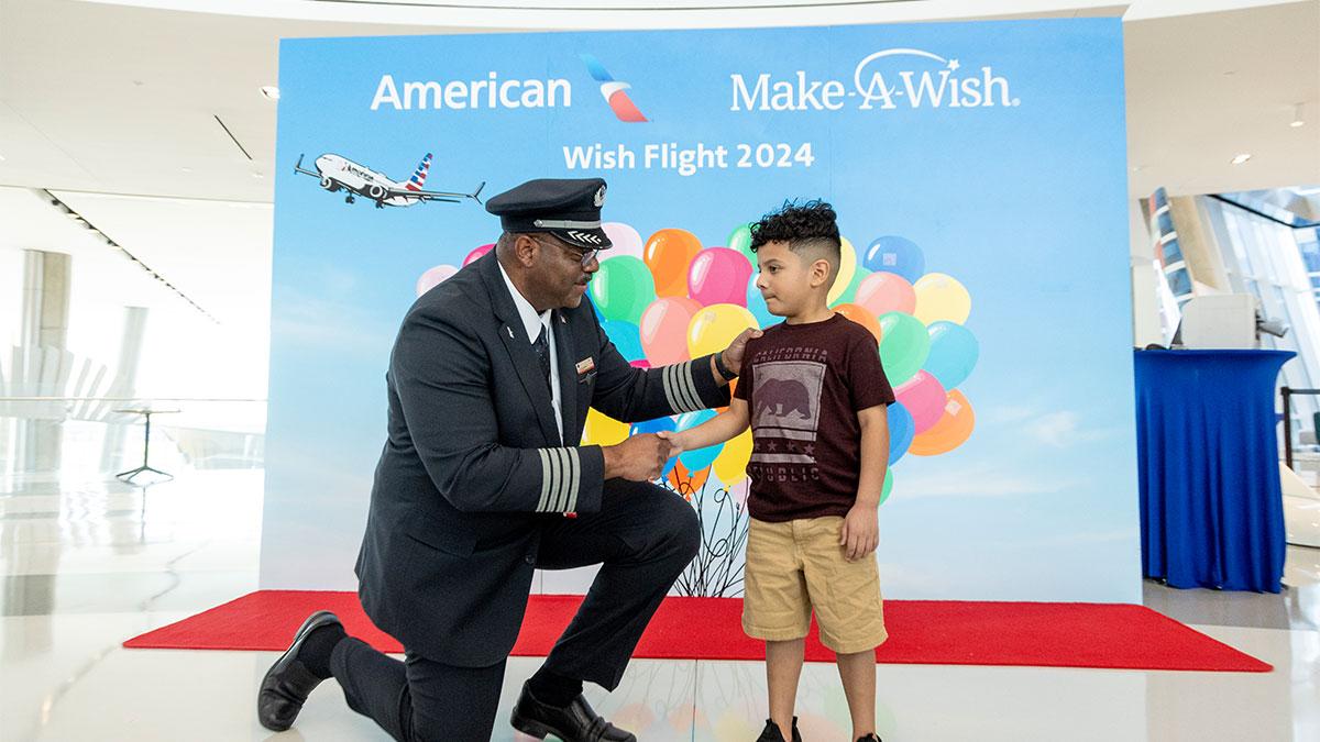 A pilot kneels and shakes the hand of a child. A poster behind them with American and Make a Wish logos, balloons.
