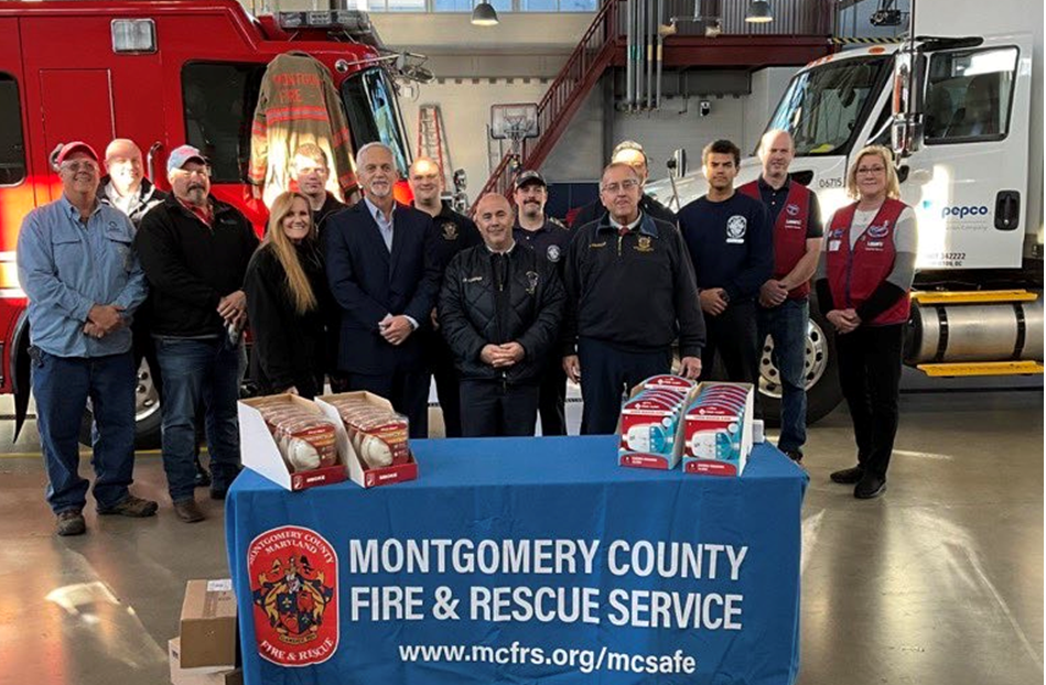 A line of people standing in front of a fire truck with a banner "Montgomery county fire and rescue service". A table with smoke detectors in front of them.