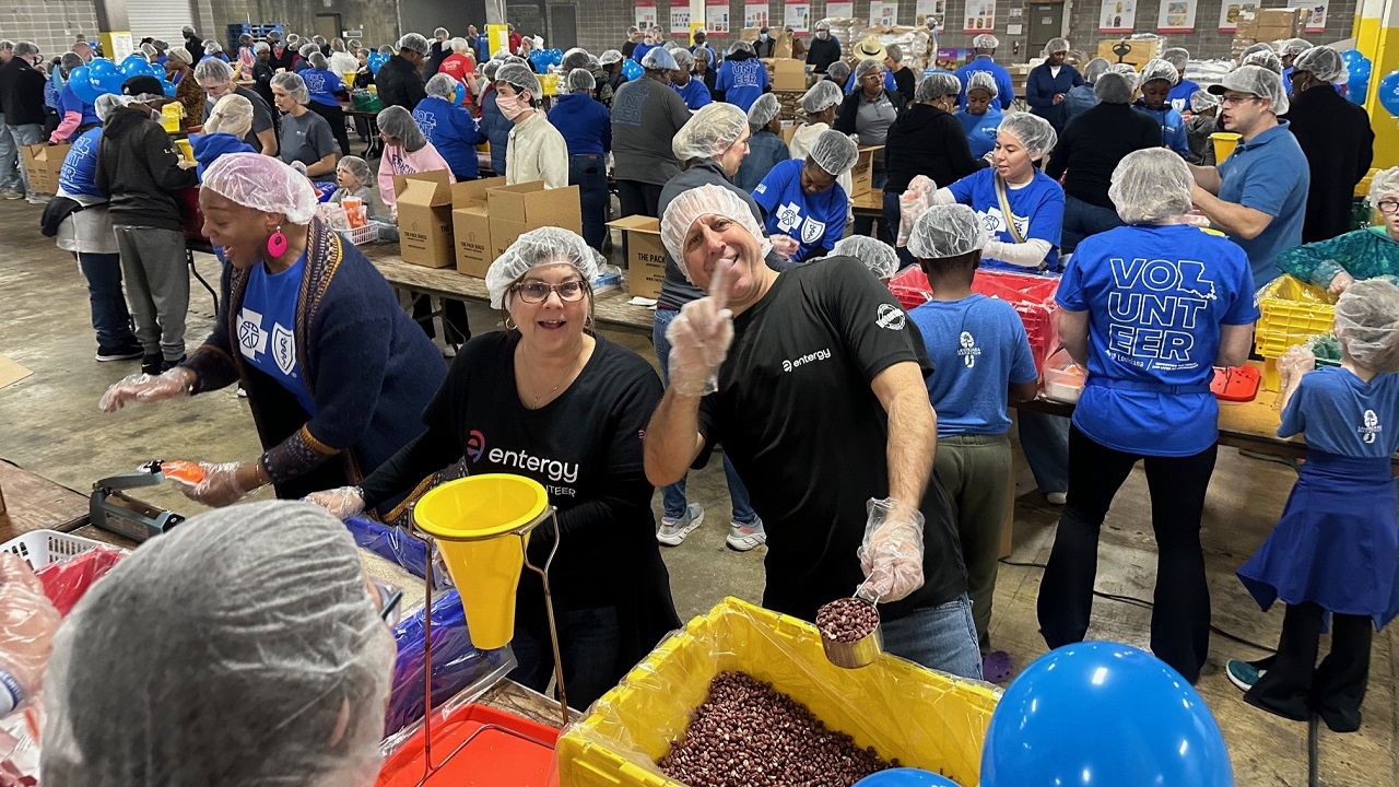 A large room full of volunteers in hair nets, packing food, two smiling to the camera