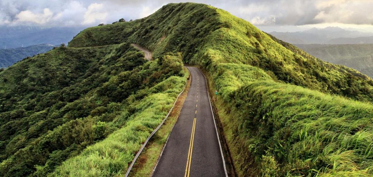 A two lane paved road leading through a greenery-covered peak. Cloudy sky in the background.