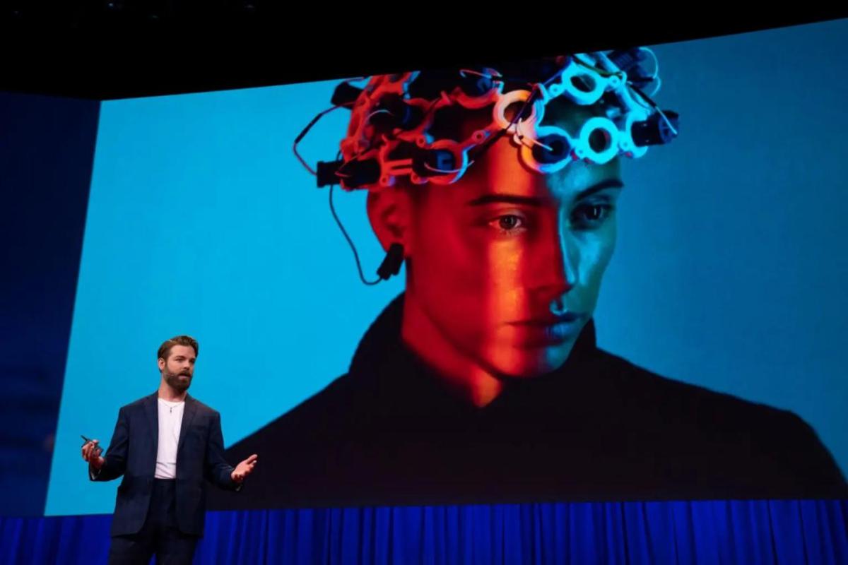 A person speaking on a stage, a digital screen showing a person with device on their head.