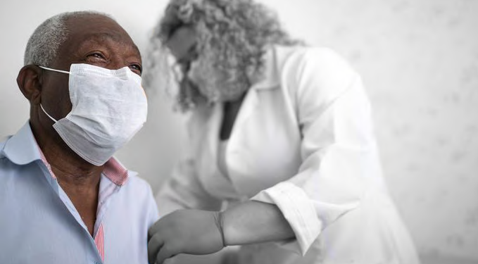 a medical professional looks at the left arm of another person. Both are wearing protective masks