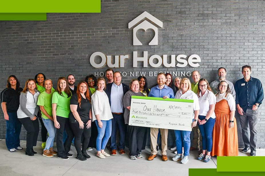 A group of people posed in front of the side of a building "Our House" sign above them. Some holding a large check made to "Our House".