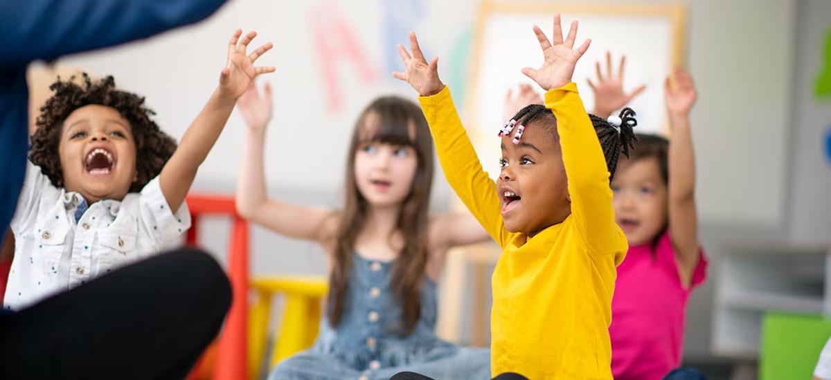 Children seated in a classroom setting, all with raised hands.