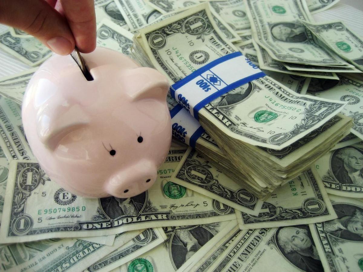 A hand putting a coin in a small piggy bank atop stacks of dollar bills.