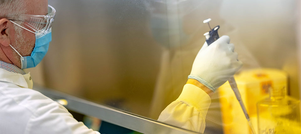 a scientist in protective gear, working behind a glass divider, using a pipette