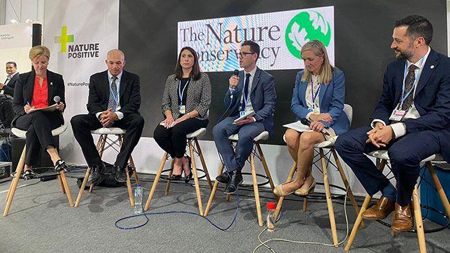 Six people sitting in tall chairs. A man in the middle speaking into a microphone. All in business attire.banner "The Nature Conservancy" behind them.