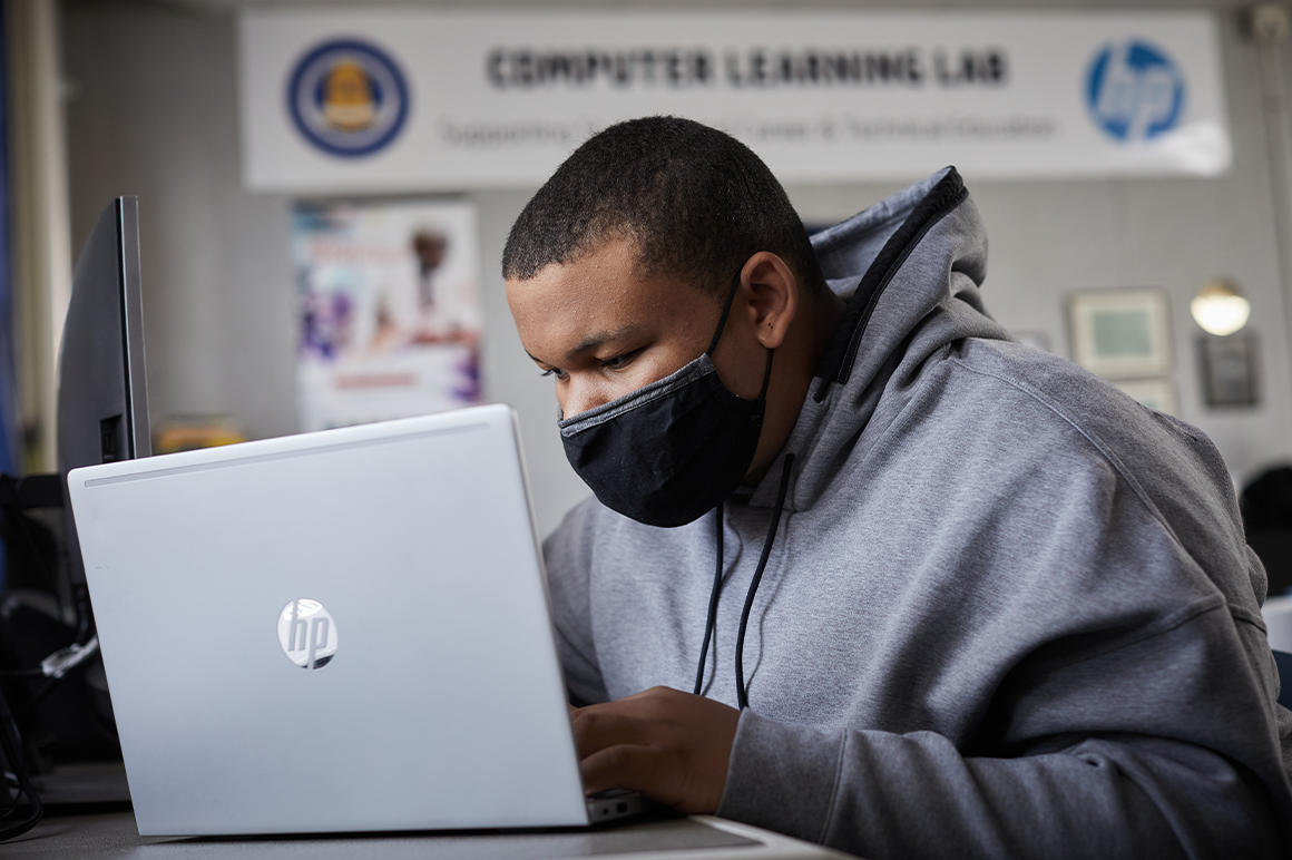 a student in a mask and gray hoodie leans close to an HP laptop screen