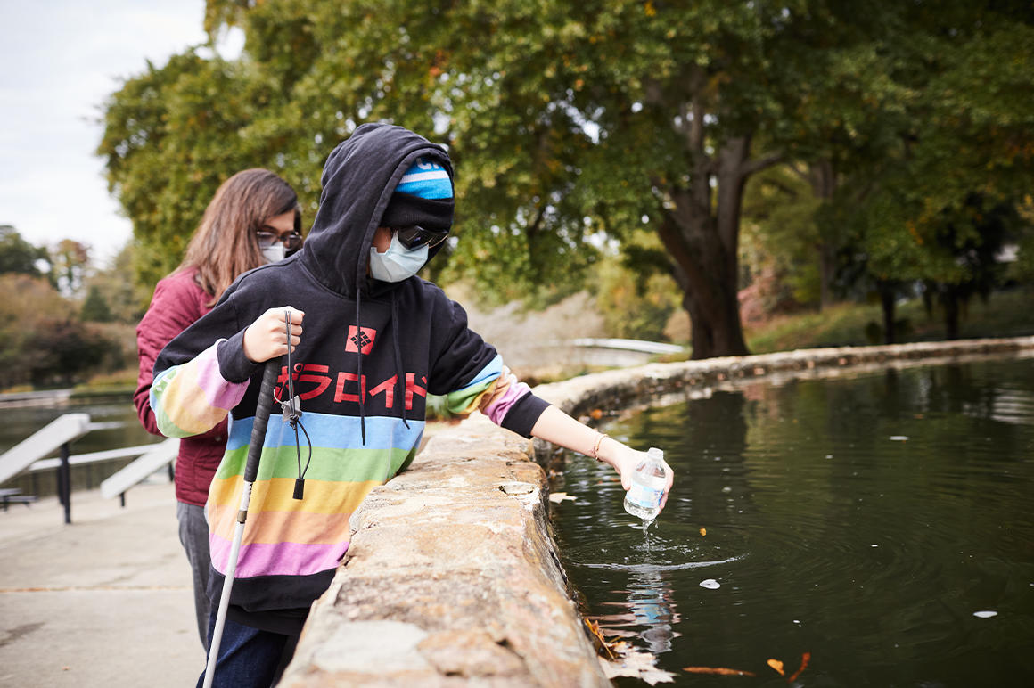 a student wearing a mask and dark sunglasses dips a water bottle into a pool
