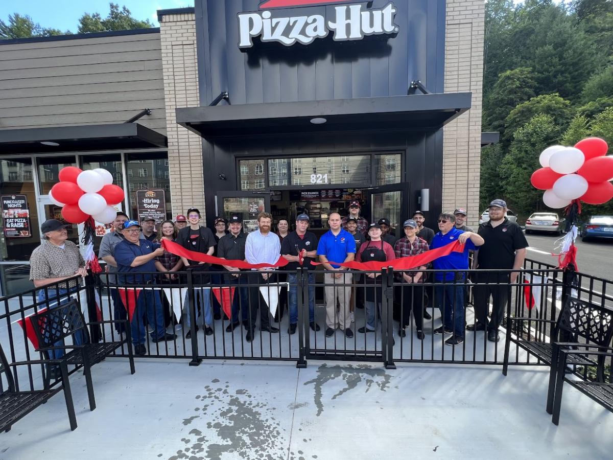 Exterior of pizza hut store, a long ribbon and red and whit balloons. A group of people cutting the ribbon.