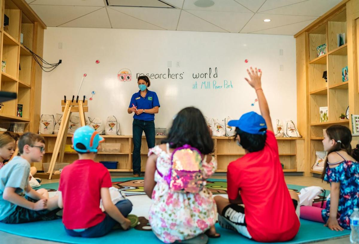 A small classroom with children seated on the floor, an adult at the front. One child has their hand up.