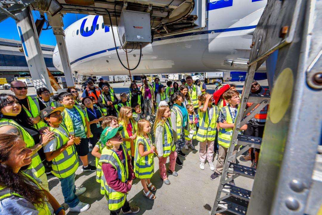 A group of kids and adults in high-vis vests looking up close at an airplane.