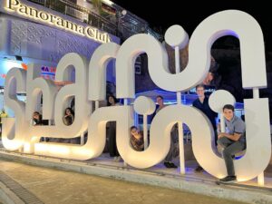 Seven people poking through a large sculpture of letters outside the "Panorama Club".