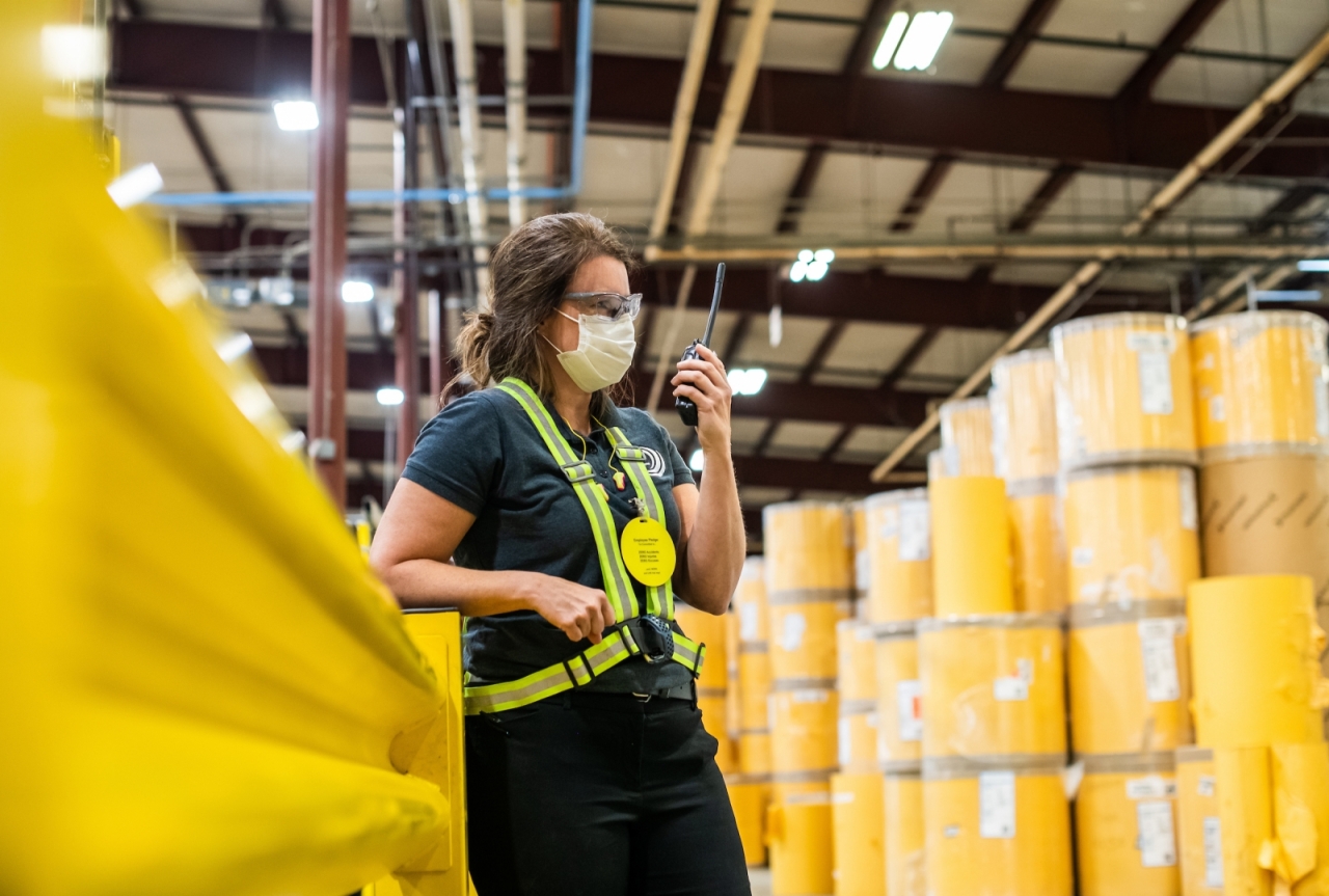 Karen Crisp in safety gear standing among yellow barrels in a factory