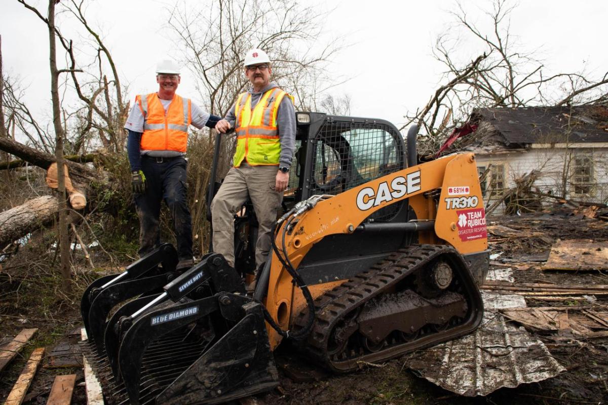 Two people in safety vests and hats standing on a CASE front-loader. Debris all around them.