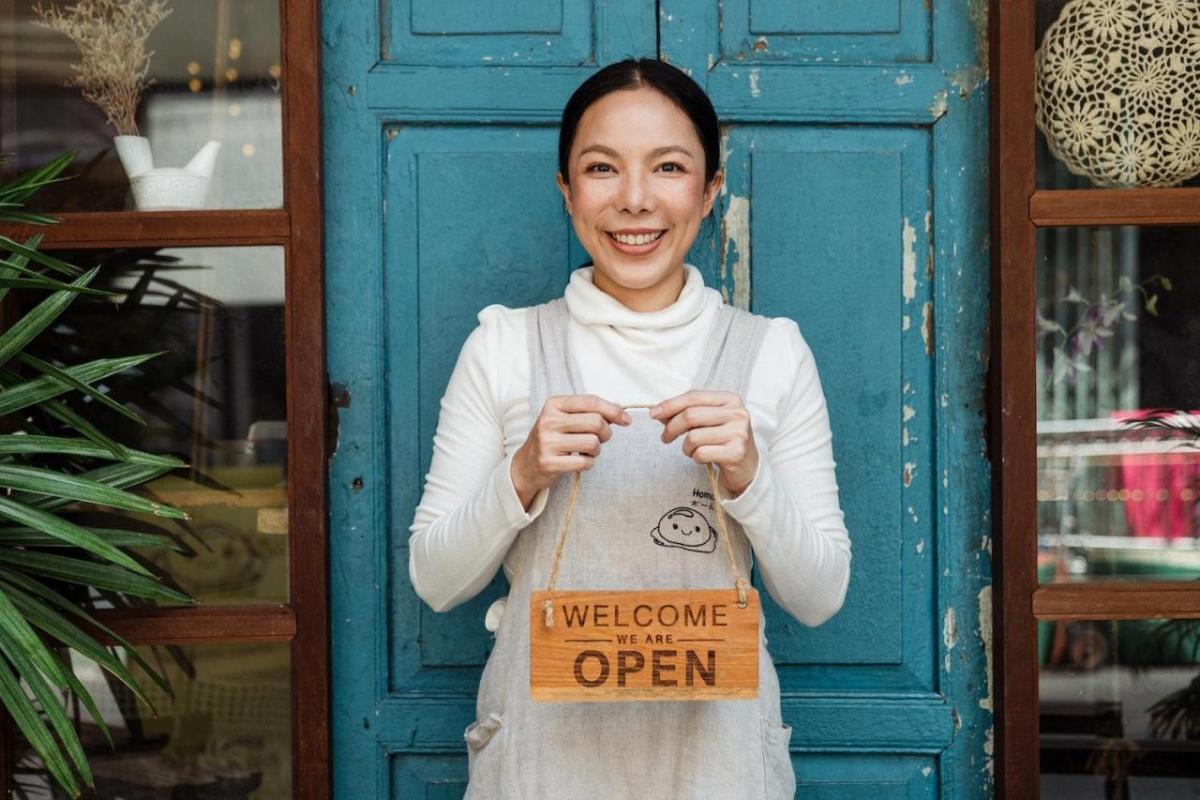A smiling person holding a wooden "Welcome we are Open" sign in front of a blue door.