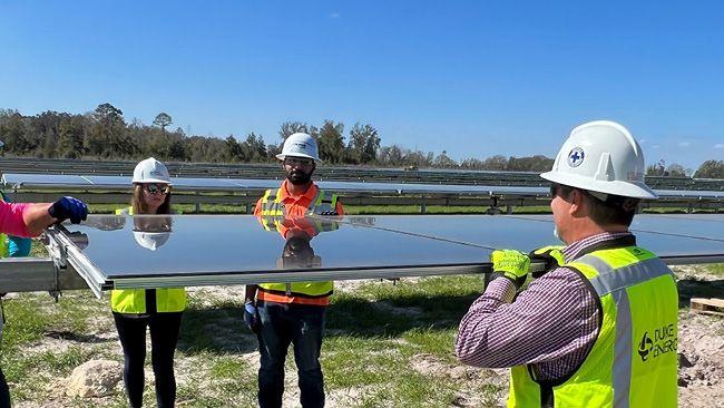 Four people installing a large solar panel in a field of other panels. All wearing safety hats, vests, gloves.