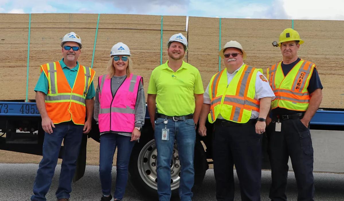 Five people in hard hats and high-vis vests in front of a trailer loaded with OSB boards.