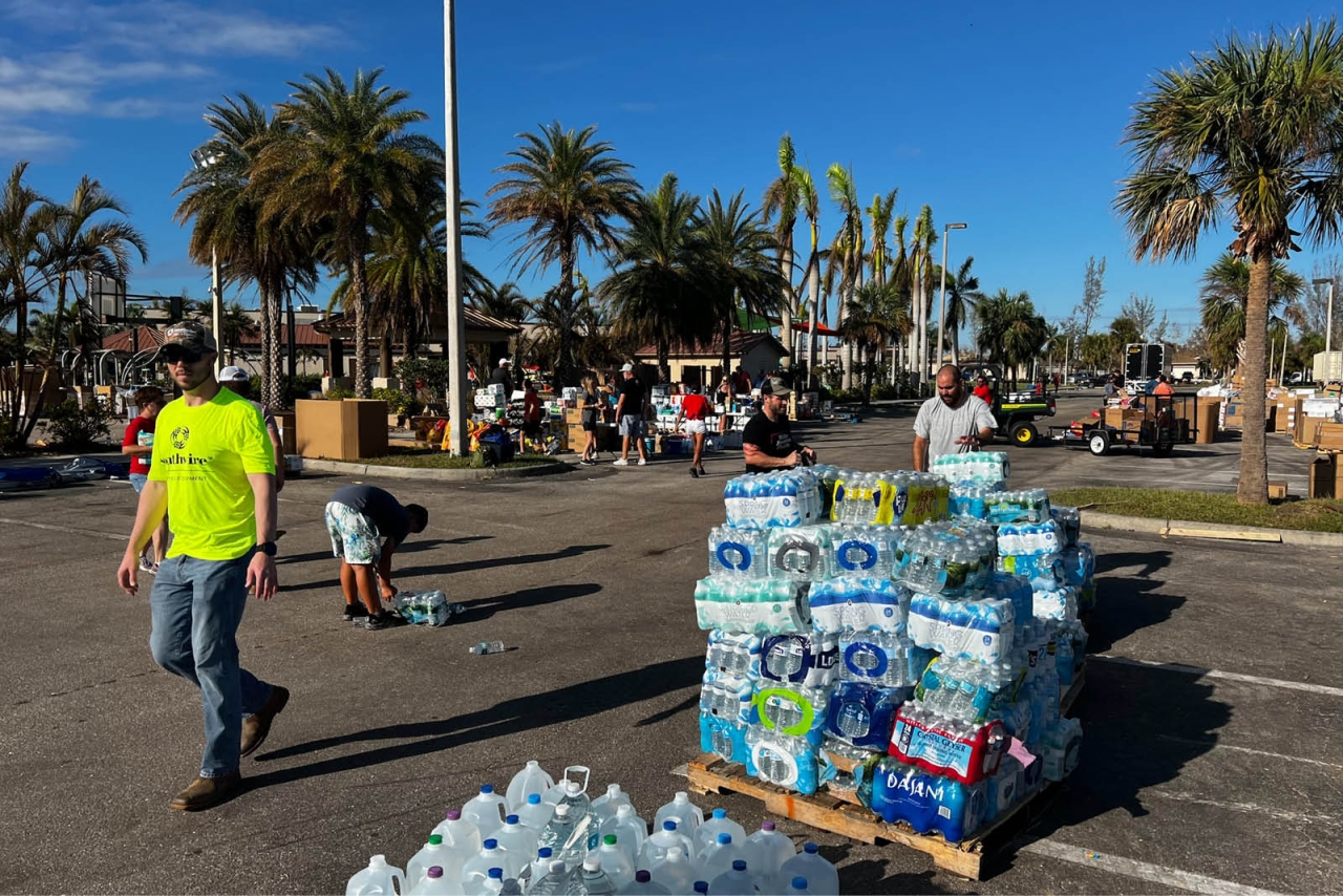 a pallet of water bottles in a parking lot, people walking around, palm trees in the distance