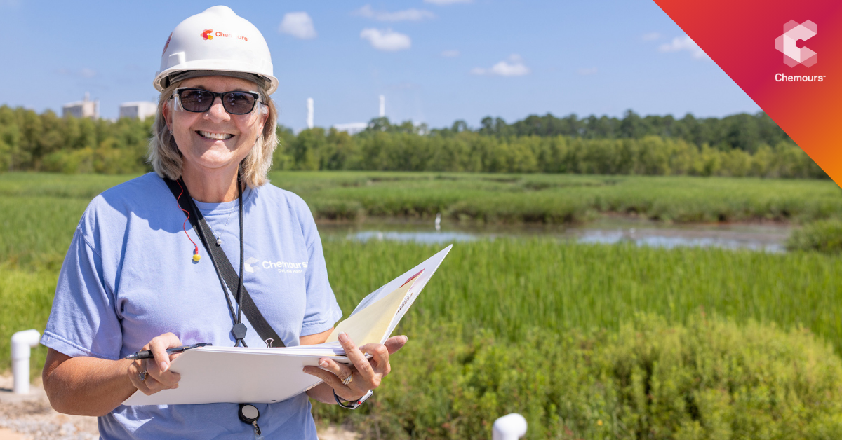 A person smiling outside, holding a notebook and wearing a hard hat