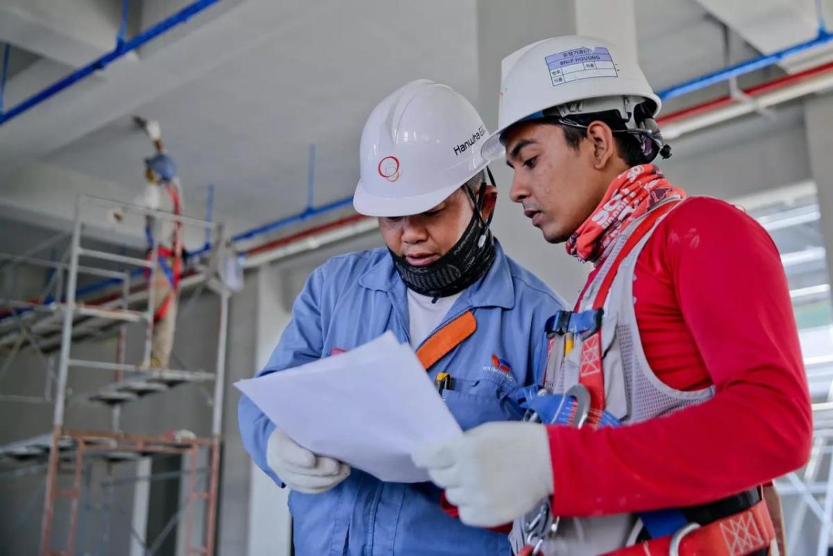 Two people in hard hats, gloves and protective masks looking at a piece of paper. A person standing on scaffolding painting the ceiling behind them.