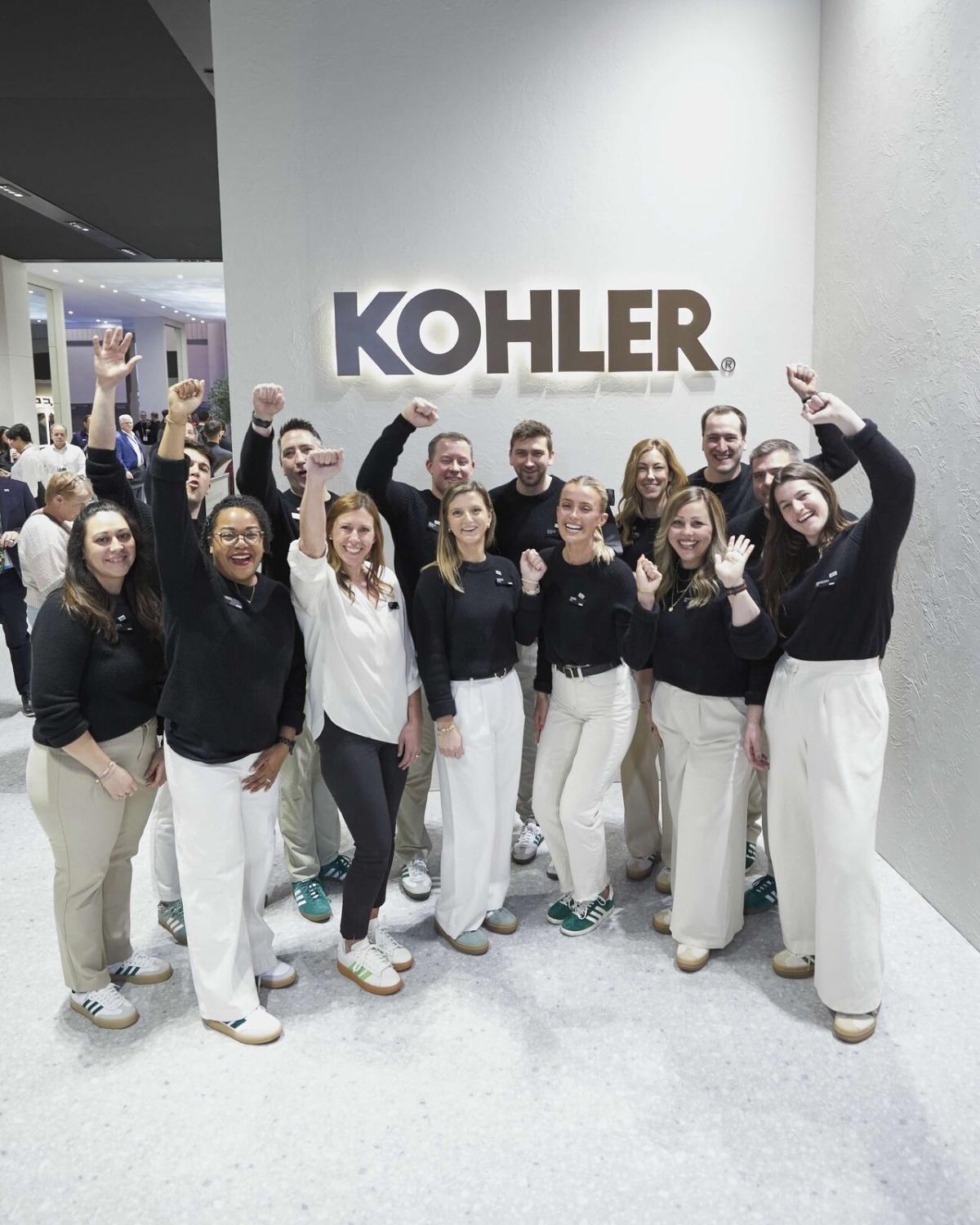 A team of people posed and cheering with arms up in front of a Kohler sign.