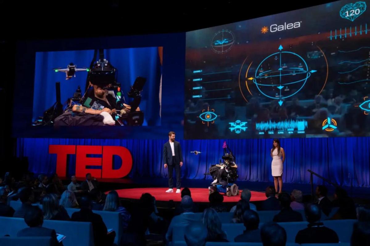 Three people on a stage "Ted" sign to the left, digital screens above.