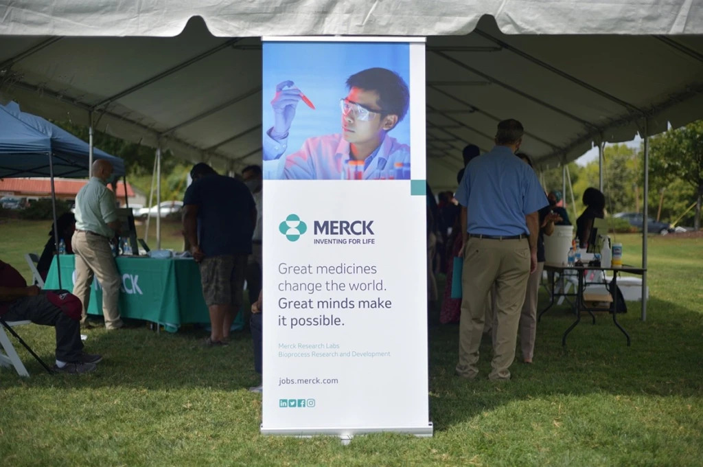 a poster board for Merck at the front of an outdoor tent, people mingling underneath or at tables