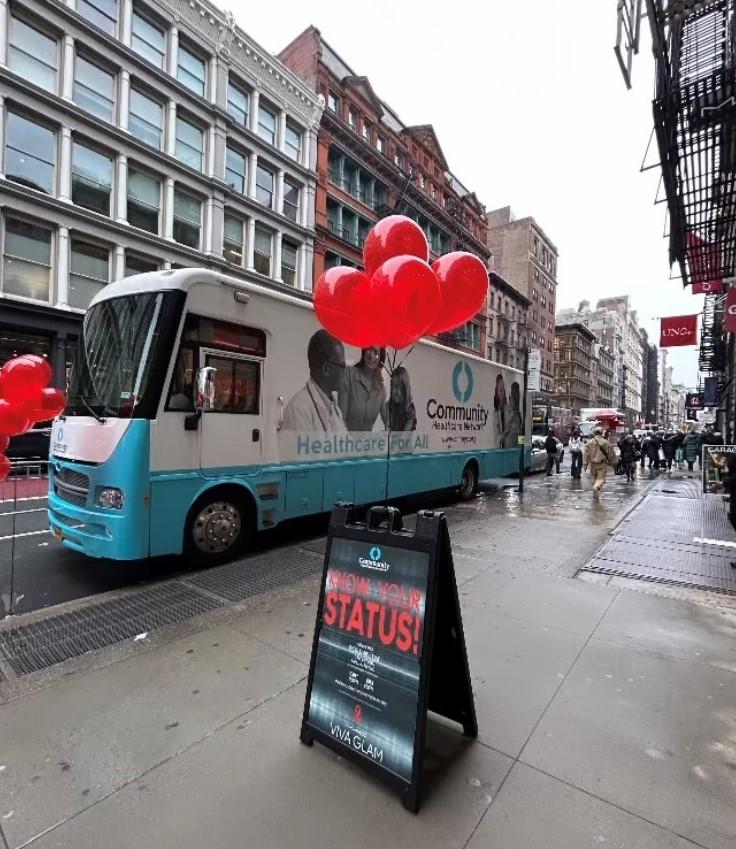 A trailer/bus parked on a street. A sign in front "Know Your Status" with red balloons.