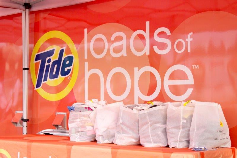 tent with banner "Tide loads of Hope". Bags of laundry on a table.