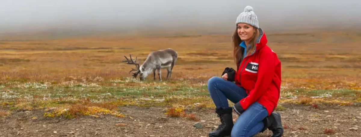 A person crouched on an open plain, a caribou in the distance