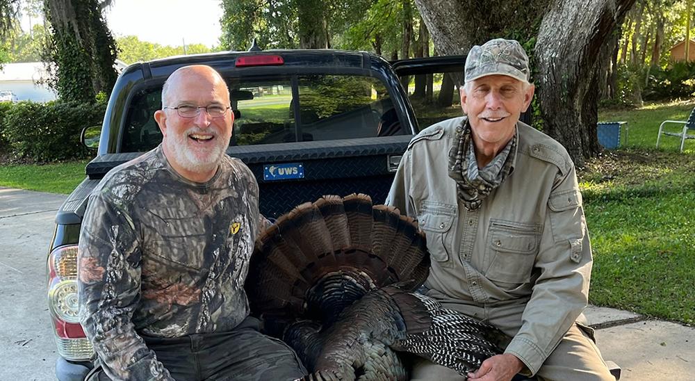 Two hunters with a turkey between them sitting in the back of a truck.