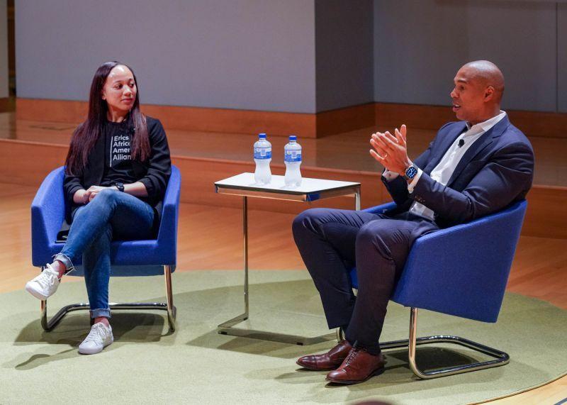 Two people seated on a lit stage, a small table between them. One speaking.