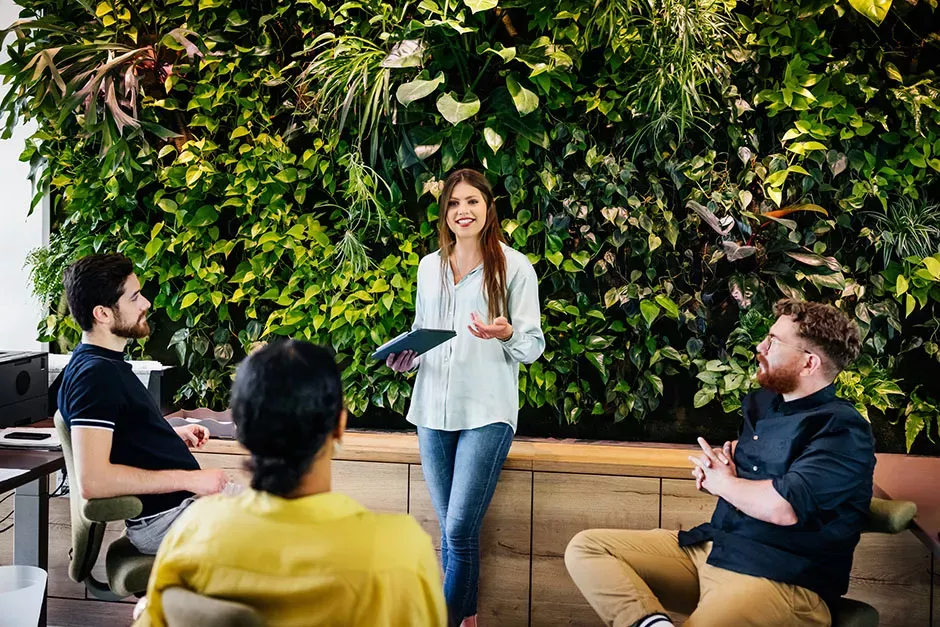 young employee in a white top stands and talks in front of a wall of green plants while coworkers listen