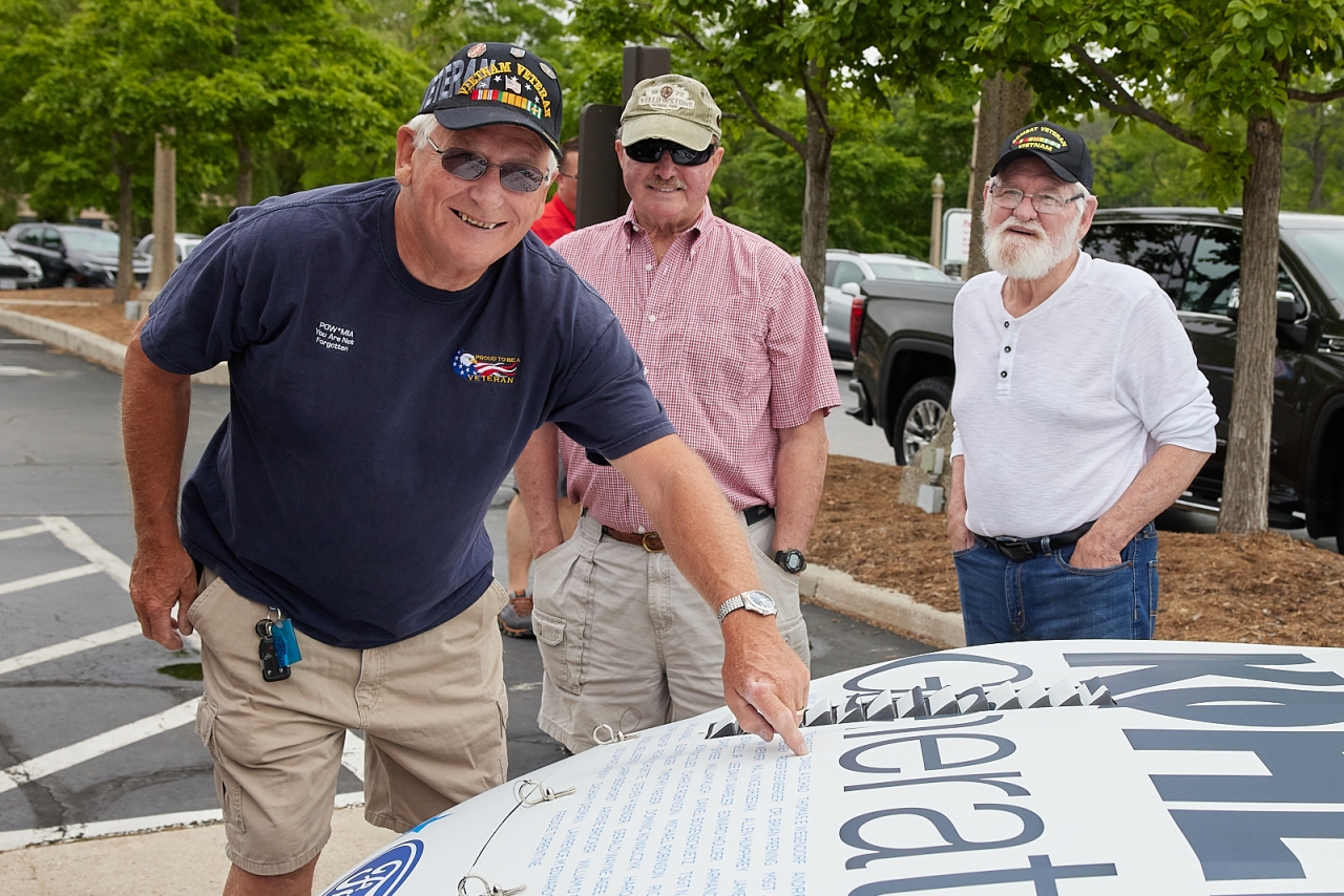 three veterans stand by a car, one points to a name on the hood