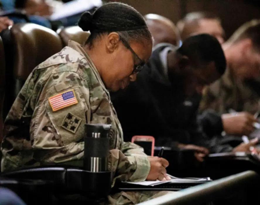 A person in military uniform seated next to others, writing in a notebook.